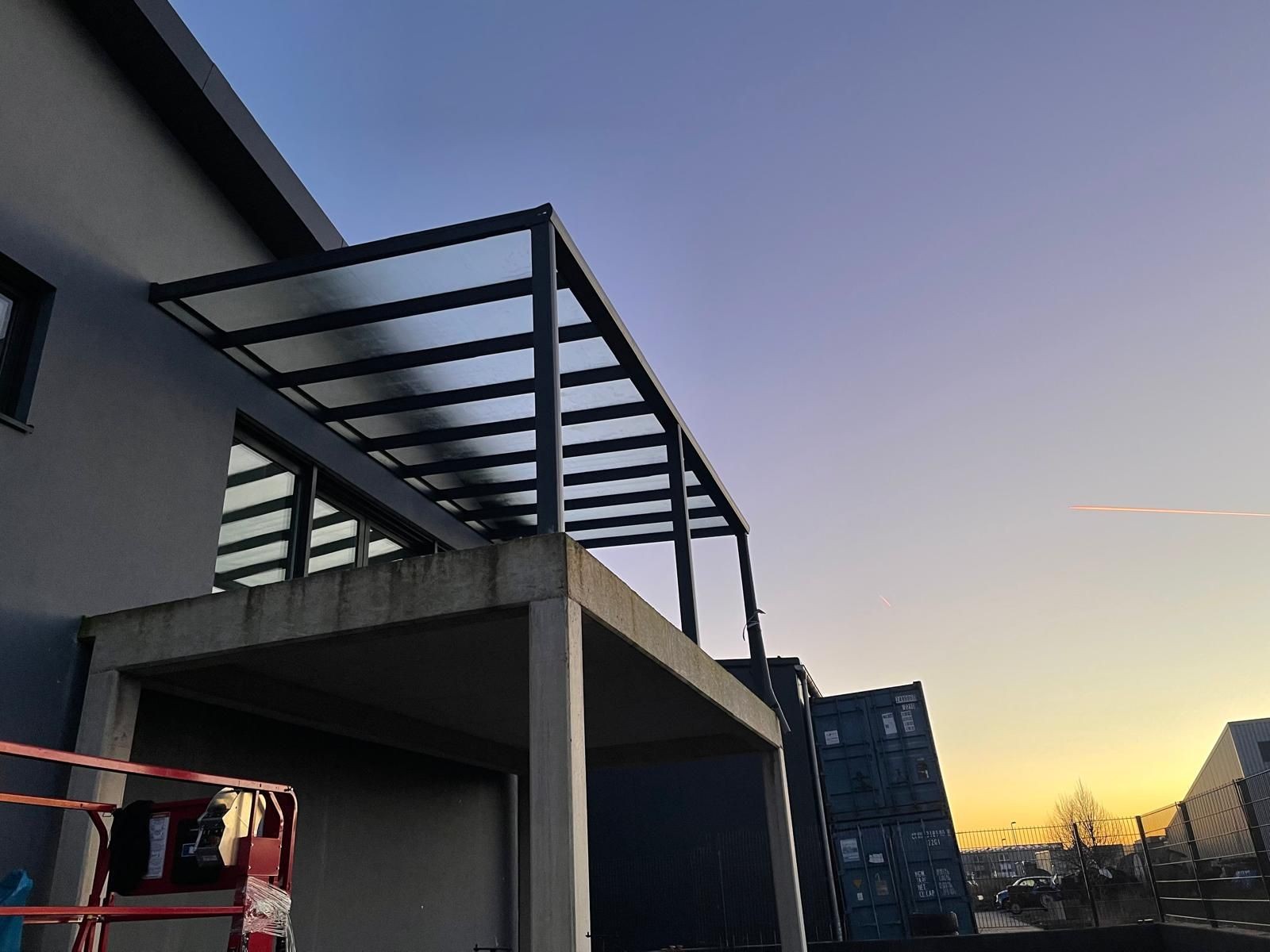 Modern building with a partially constructed glass roof structure at sunset with industrial containers in the background.